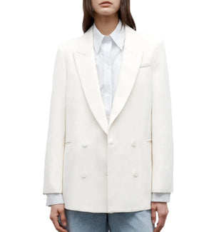 A woman wears the TWP Tabitha Blazer—a white cotton-linen, double-breasted jacket—over a light collared shirt and blue jeans, standing against a white background.