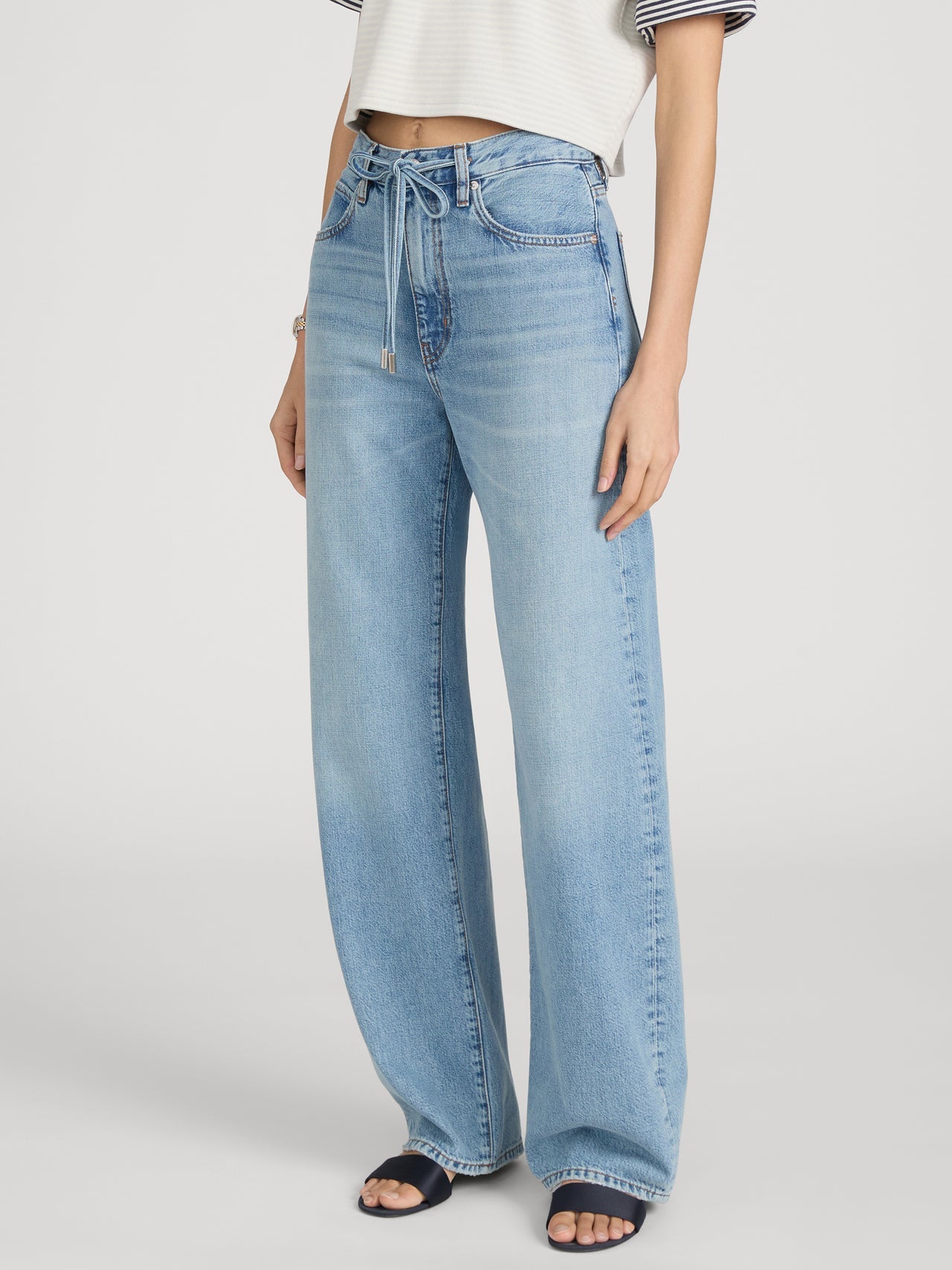 A person wears Frame's The Off Duty Drawstring Bow high-waisted, light blue barrel silhouette jeans with a drawstring belt, styled with a cropped striped top and dark sandals.