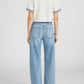 A person stands facing away in Frame’s The Off Duty Drawstring Bow white short-sleeve top with striped cuffs, paired with lightweight denim high-rise barrel-leg jeans and black open-toe heels against a plain background.