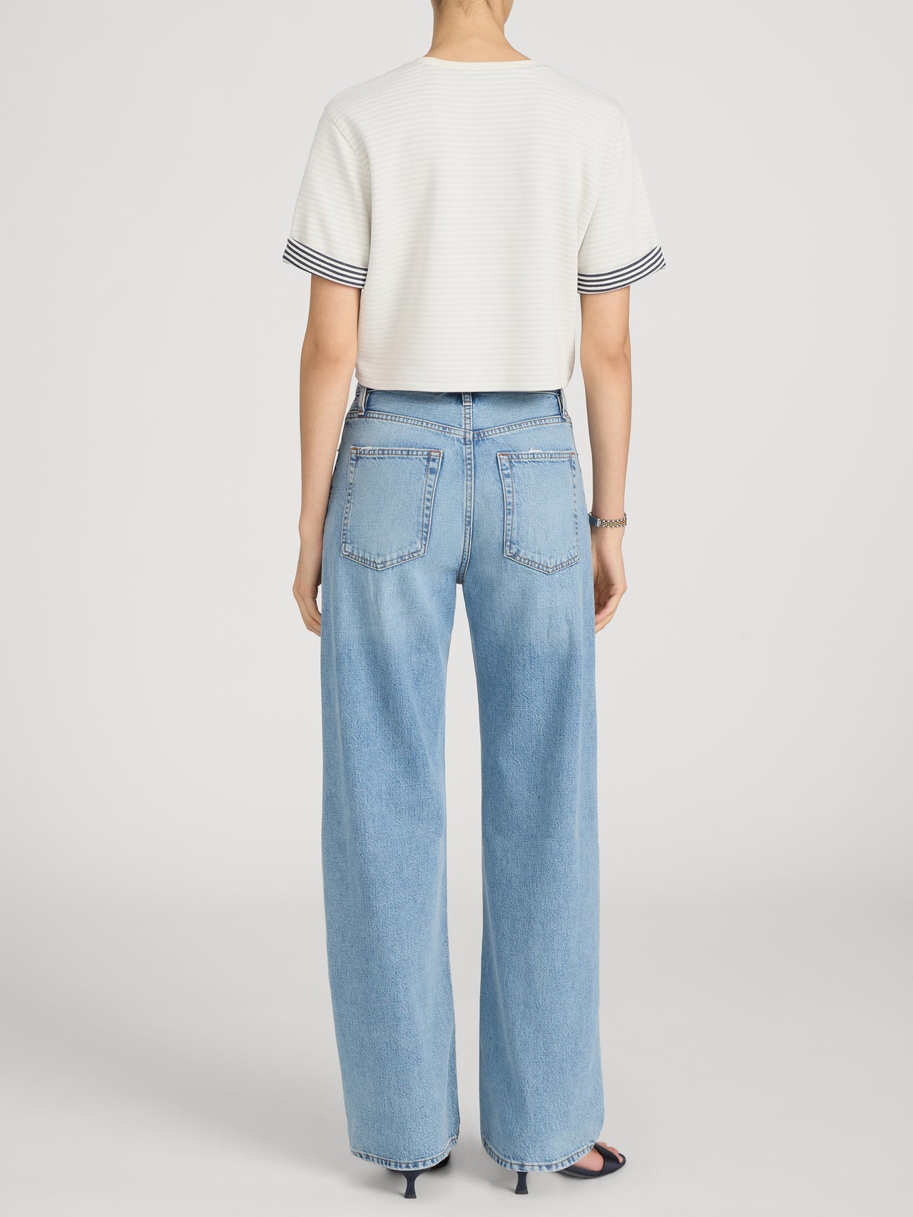 A person stands facing away in Frame’s The Off Duty Drawstring Bow white short-sleeve top with striped cuffs, paired with lightweight denim high-rise barrel-leg jeans and black open-toe heels against a plain background.