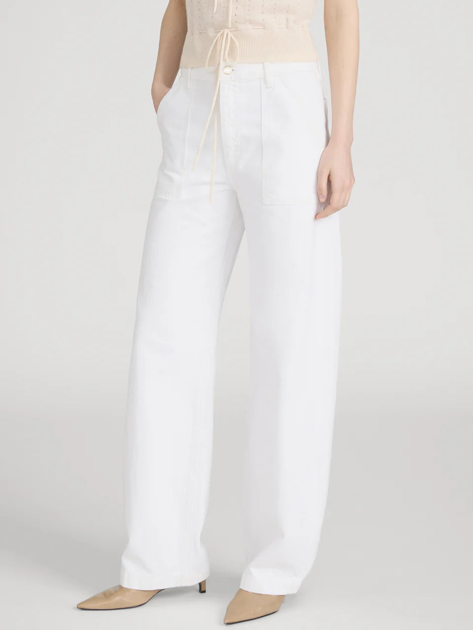 Wearing Frame’s The Bow Cargo high-rise barrel jeans with front cargo pockets, a light sleeveless top, and beige pointed-toe heels, a person stands against a plain background for a modern utilitarian look.