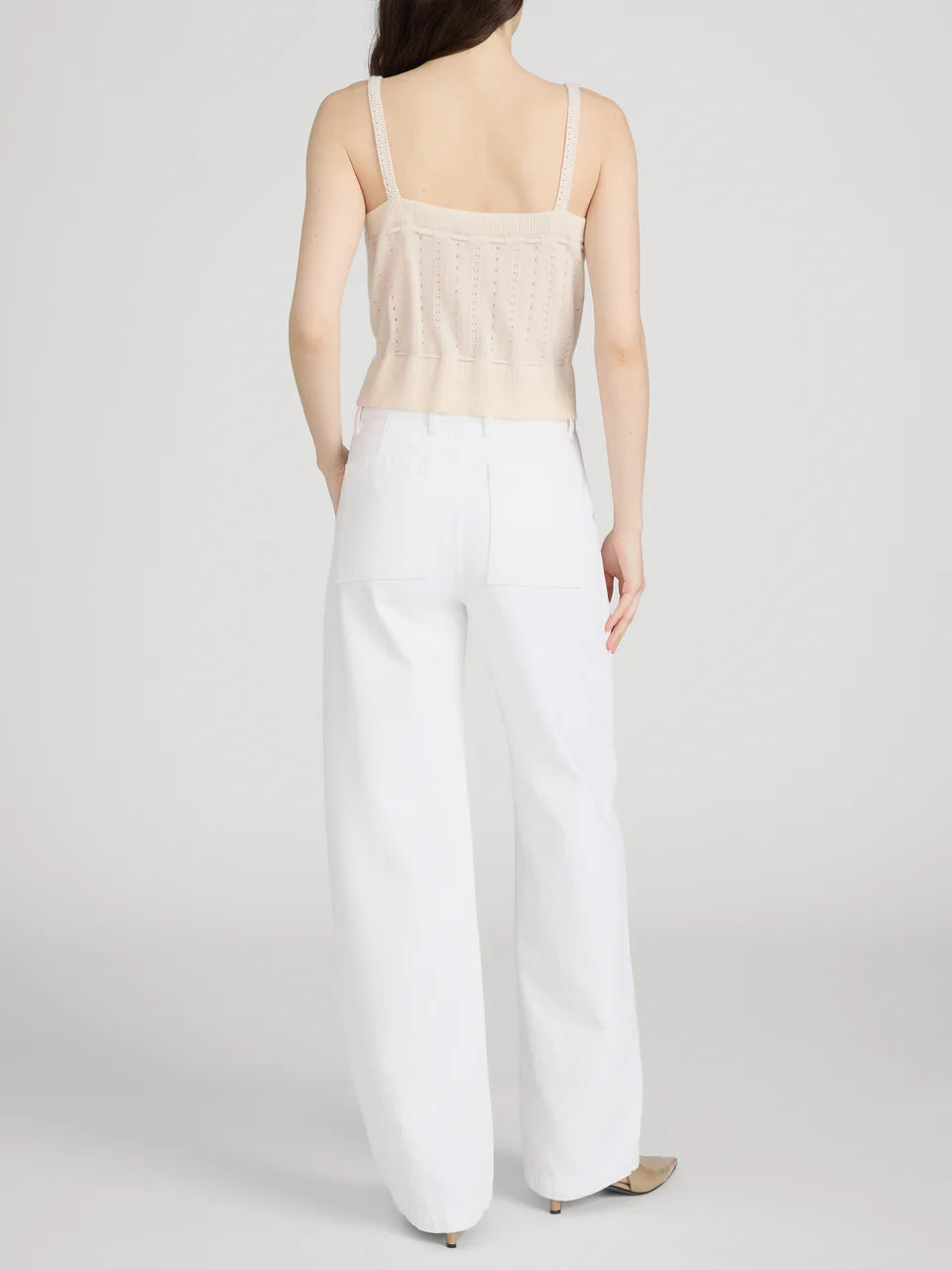 A woman stands facing away, wearing Frame's The Bow Cargo jeans paired with a beige sleeveless knit top and heeled shoes, set against a plain light background.