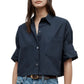 A woman wears the TWP Next Ex Shirt—a navy blue vintage-washed poplin button-up with rolled sleeves, front pocket, and boxy silhouette—paired with blue jeans and a brown belt.