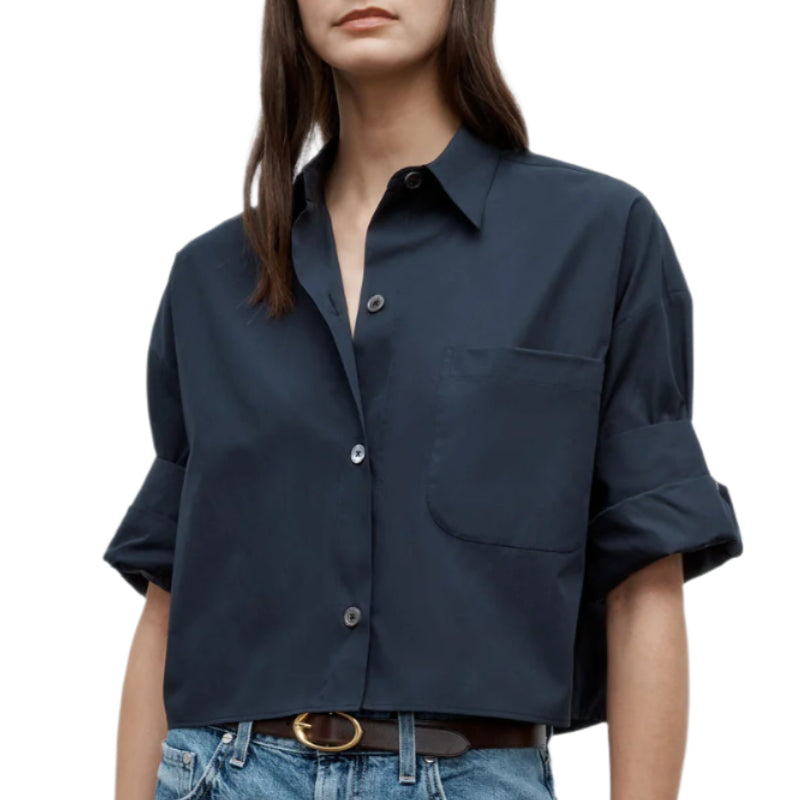 A woman wears the TWP Next Ex Shirt—a navy blue vintage-washed poplin button-up with rolled sleeves, front pocket, and boxy silhouette—paired with blue jeans and a brown belt.