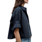 A woman wears the TWP Next Ex Shirt, a vintage washed poplin short-sleeve button-up with rolled cuffs, paired with light blue jeans, shown from the side against a white background.