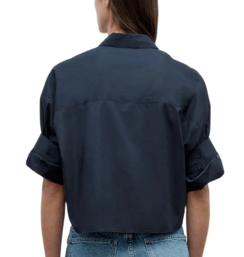 A person with long brown hair is seen from the back wearing the TWP Next Ex Shirt, a dark blue vintage-washed poplin cropped shirt with a boxy silhouette, paired with blue jeans, set against a plain white background.