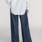 A person stands with their back to the camera, wearing a loose-fitting, light blue striped shirt paired with Frame’s The Offshore Pant—lightweight, wide-leg pants featuring a drawstring waistband.