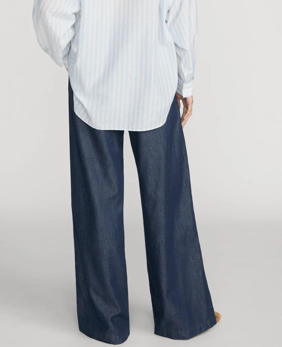 A person stands with their back to the camera, wearing a loose-fitting, light blue striped shirt paired with Frame’s The Offshore Pant—lightweight, wide-leg pants featuring a drawstring waistband.