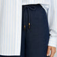 A person wears a light blue and white striped button-up shirt tucked into Frame’s The Offshore Pant, a pair of high-waisted, lightweight wide leg pants featuring a drawstring waistband with gold tips.