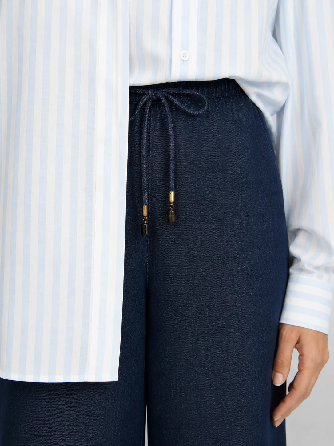 A person wears a light blue and white striped button-up shirt tucked into Frame’s The Offshore Pant, a pair of high-waisted, lightweight wide leg pants featuring a drawstring waistband with gold tips.