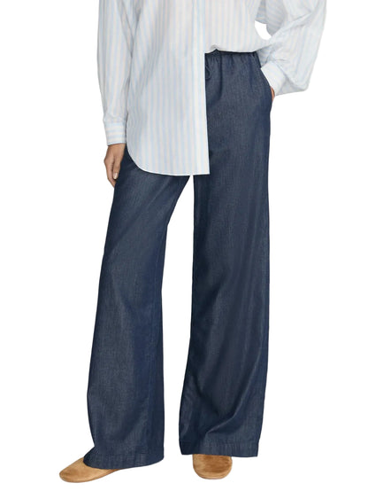 A person is wearing a blue and white striped shirt, tan shoes, and The Offshore Pant by Frame—lightweight, wide-leg dark blue pants with a drawstring waistband—while standing against a plain background.