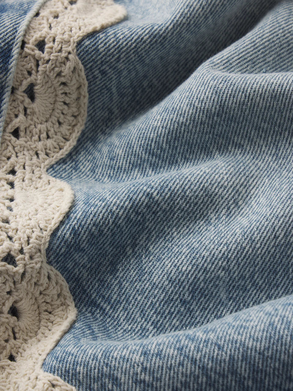 Close-up of light blue denim from Frame’s The Crochet Bow Crop, detailed with a delicate cream crochet trim along one edge.