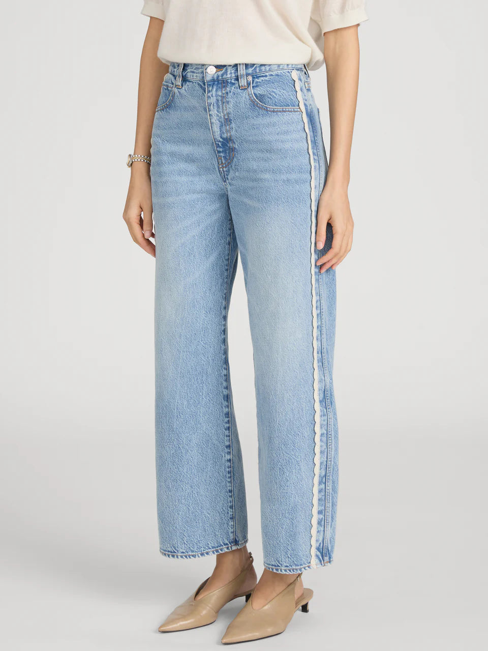 A person wears The Crochet Bow Crop by Frame with light wash, high-rise wide-leg jeans featuring a white side stripe and beige pointed-toe shoes, standing against a plain background.