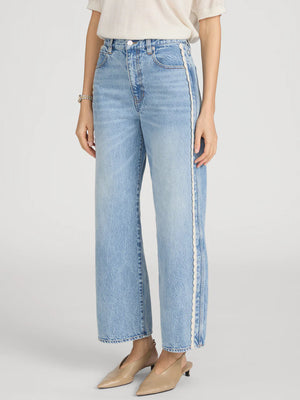 A person wears The Crochet Bow Crop by Frame with light wash, high-rise wide-leg jeans featuring a white side stripe and beige pointed-toe shoes, standing against a plain background.