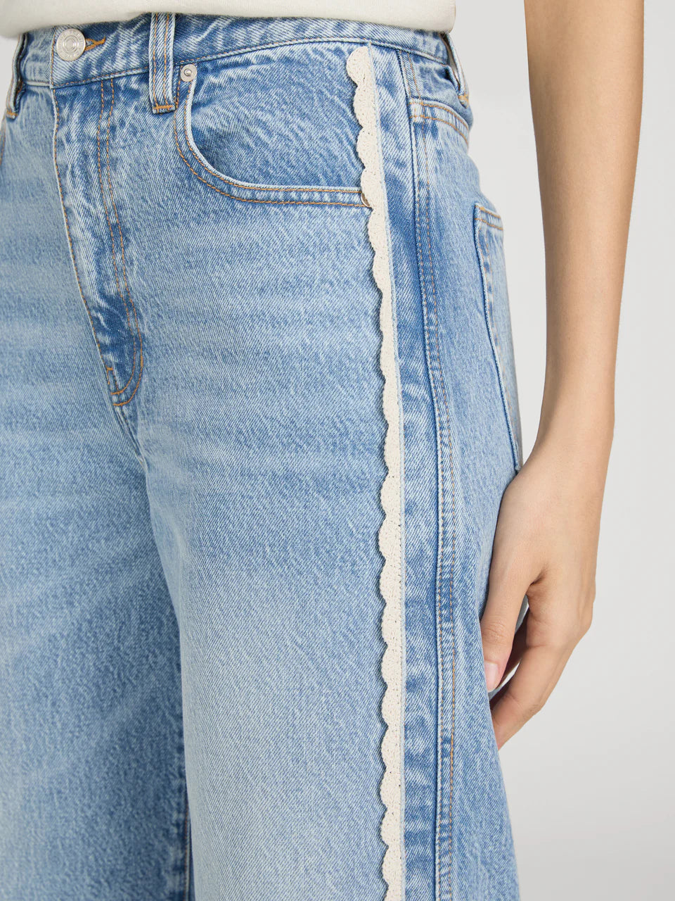 Close-up of a person wearing Frame’s The Crochet Bow Crop jeans in light blue denim, featuring scalloped white crochet trim along the side seam; their hand rests at their side.
