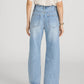 A person is shown from the back wearing Frame’s The Crochet Bow Crop—a short-sleeve beige top with crochet trim—paired with light blue high-rise, wide-leg Bow Crop jeans and beige low-heeled shoes.