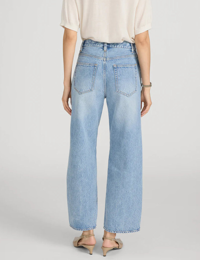 A person is shown from the back wearing Frame’s The Crochet Bow Crop—a short-sleeve beige top with crochet trim—paired with light blue high-rise, wide-leg Bow Crop jeans and beige low-heeled shoes.
