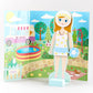 Elsie Magnetic Dress Up Character by Floss and Rock stands in front of a colorful park scene featuring an ice cream truck, pool, picnic, swing, and trees, wearing a stylish white and yellow outfit.