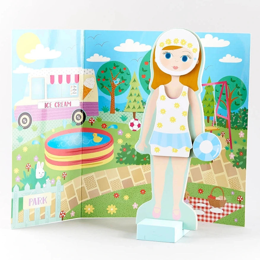 Elsie Magnetic Dress Up Character by Floss and Rock stands in front of a colorful park scene featuring an ice cream truck, pool, picnic, swing, and trees, wearing a stylish white and yellow outfit.