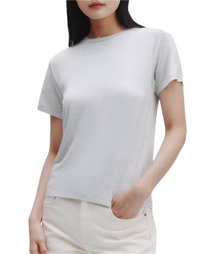A woman stands against a white background, wearing the TWP His Tee—an ultra-soft cotton crewneck t-shirt in light gray—and off-white pleated pants, with one hand in her pocket.
