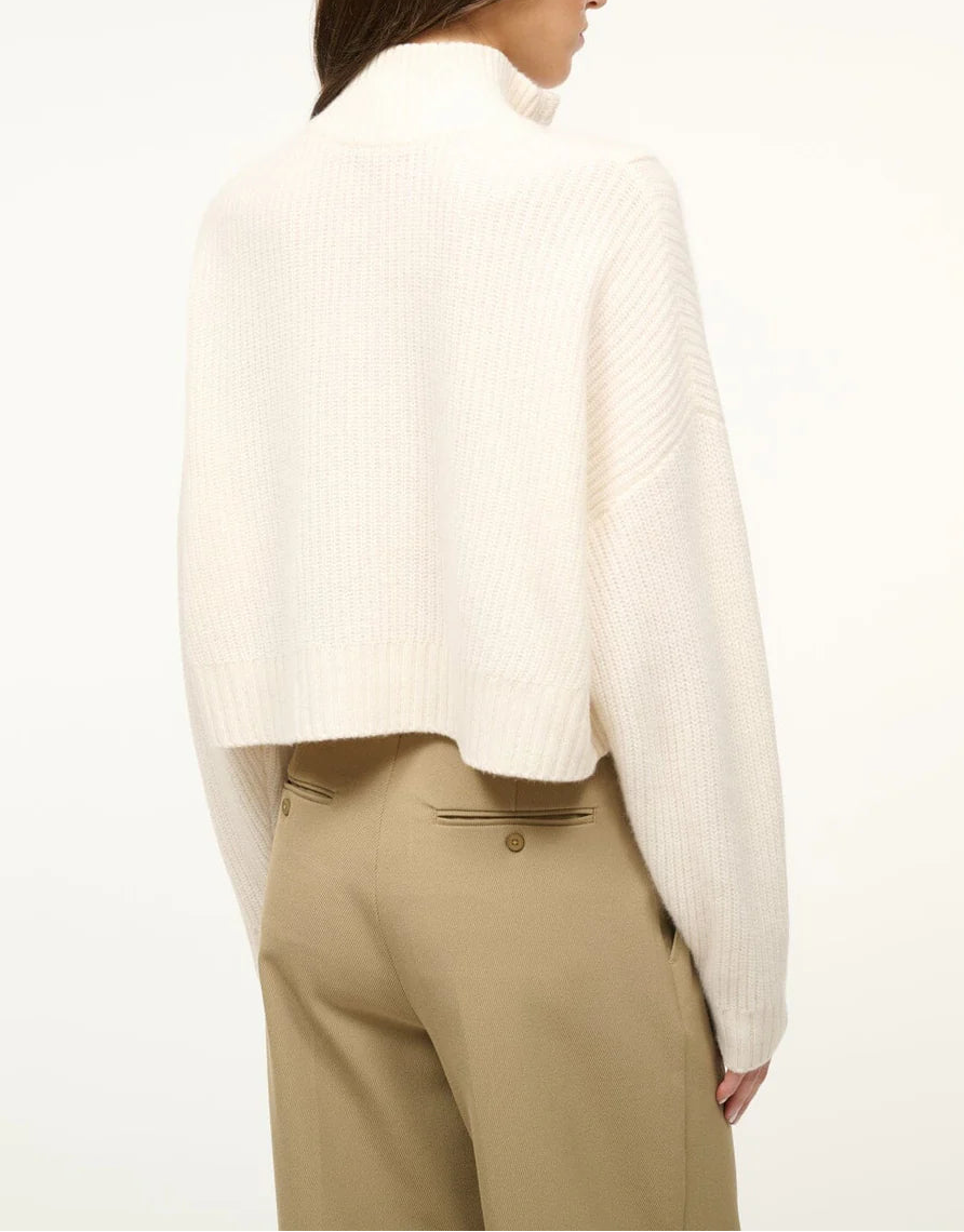 A person shown from the back wearing the Staud Cropped Hampton Cashmere Sweater in white with long sleeves, paired with high-waisted beige trousers.