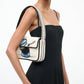 A woman in a black spaghetti strap dress carries the Staud Tommy Beaded Bag, a cream-colored vegan leather shoulder bag featuring a blue and black bowl and spoon graphic.