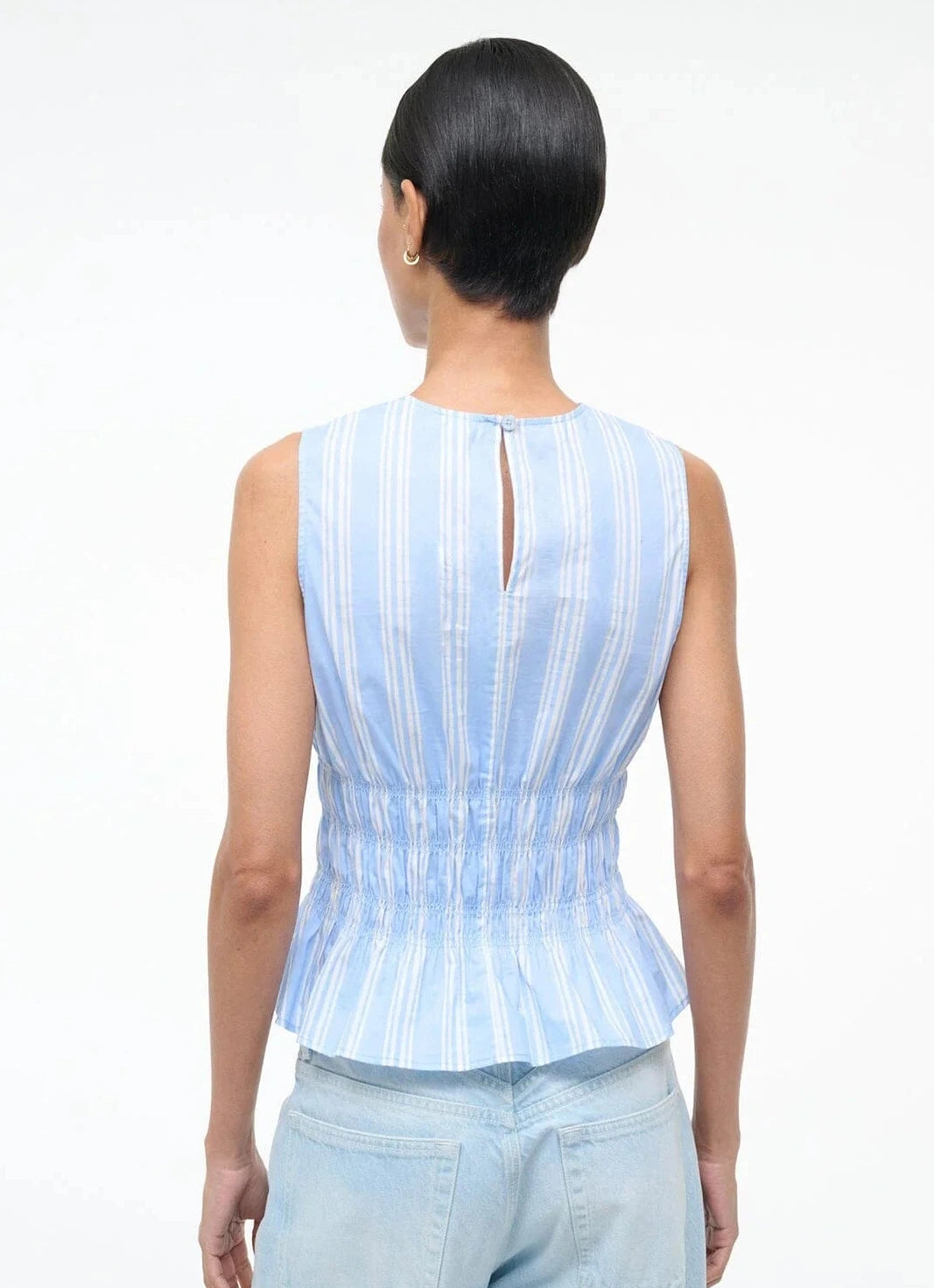 A person photographed from behind wears the Staud Culver Top—a sleeveless, light blue and white striped blouse with a keyhole button back and fitted silhouette—paired with light blue jeans.