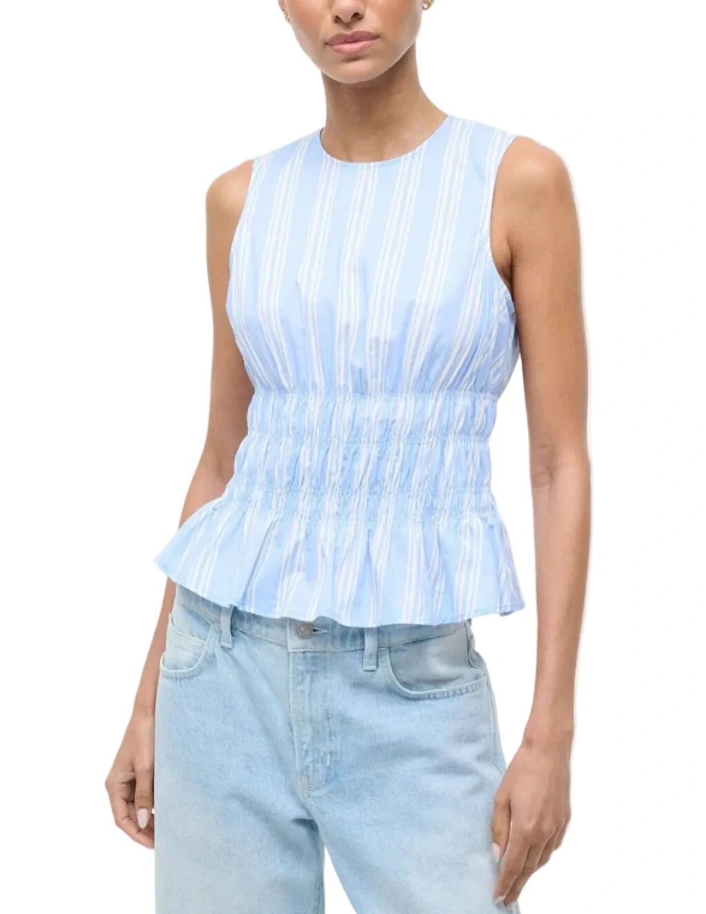 A woman wears the Staud Culver Top—a sleeveless, light blue and white striped peplum top with waist ruching—paired with light blue jeans.