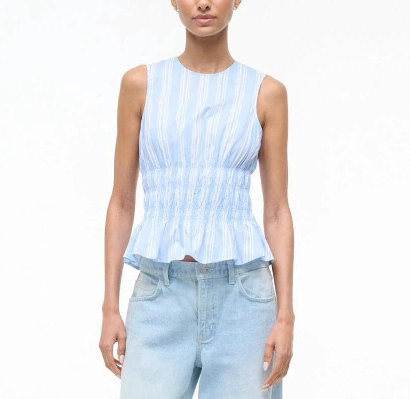 A person stands against a plain white background wearing the Staud Culver Top, a sleeveless blue and white striped fitted top with waist ruching, paired with light blue jeans.