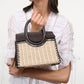 A woman in a white blouse holds the Maude Mini Wicker Carryall by Staud—a structured handbag with a woven straw body and dark leather accents and handles.