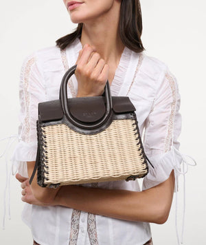 A woman in a white blouse holds the Maude Mini Wicker Carryall by Staud—a structured handbag with a woven straw body and dark leather accents and handles.