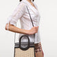 A woman in a white blouse and plaid shorts carries the Staud Maude Mini Wicker Carryall, a structured handheld bag with black leather handles and a versatile shoulder strap.