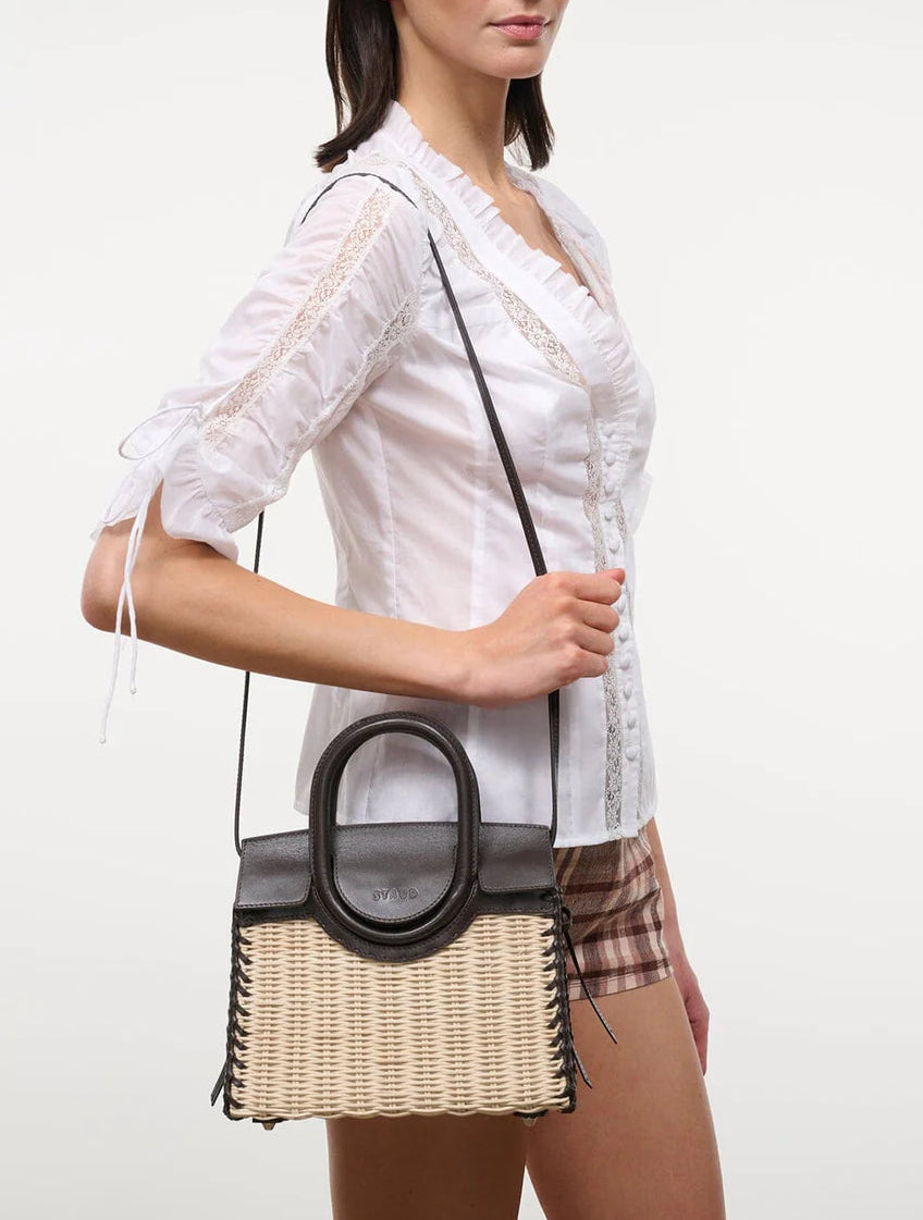 A woman in a white blouse and plaid shorts carries the Staud Maude Mini Wicker Carryall, a structured handheld bag with black leather handles and a versatile shoulder strap.