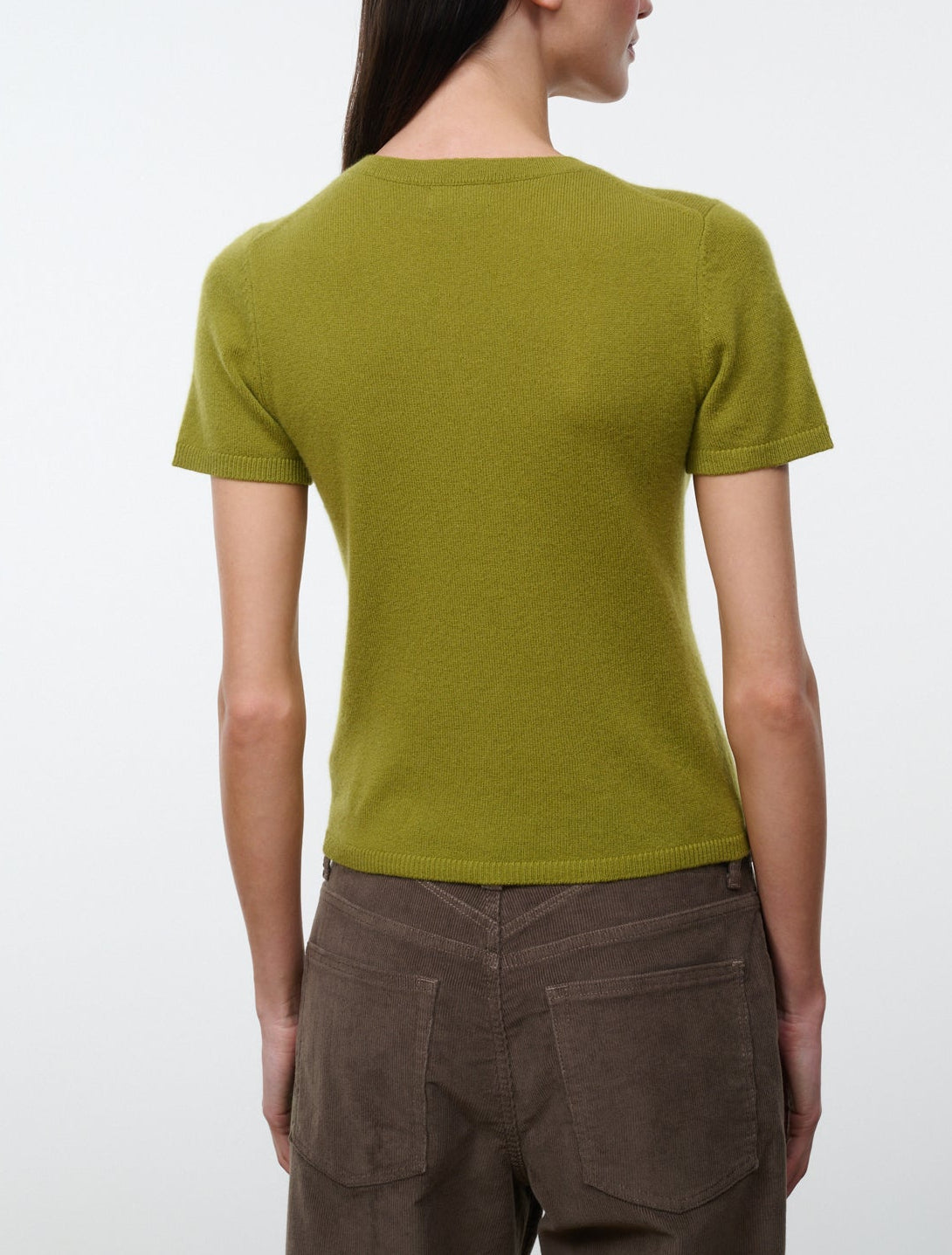 A person stands facing away against a plain light background, wearing the Staud Easy Cashmere Tee—a short-sleeved, olive green cashmere top with a crew neckline—paired with brown pants.