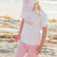 A young girl stands on a sandy beach wearing light pink pants, a white shirt, and the Bits & Bows Big Sis Bow Baseball Hat, both featuring "big sister" in pink letters—perfect as a big sister gift.