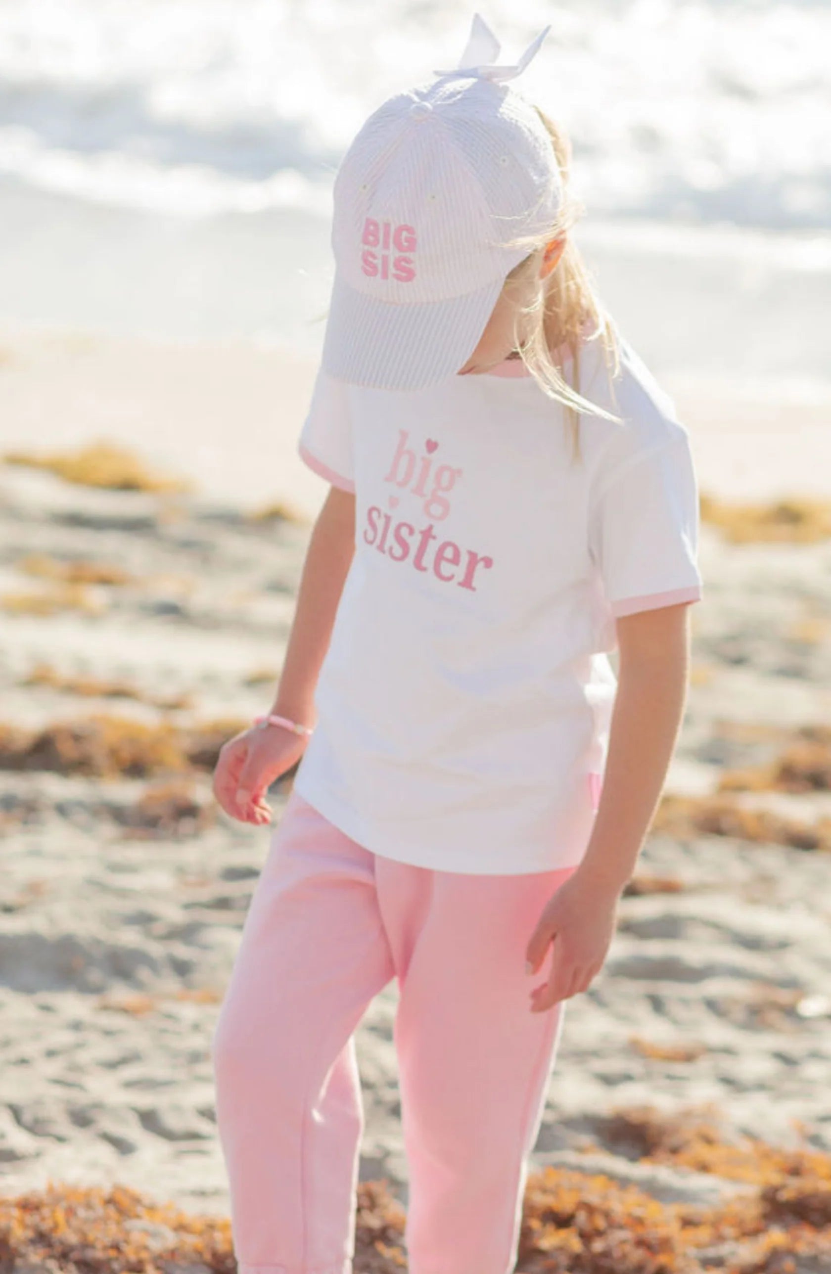 A young girl stands on a sandy beach wearing light pink pants, a white shirt, and the Bits & Bows Big Sis Bow Baseball Hat, both featuring "big sister" in pink letters—perfect as a big sister gift.
