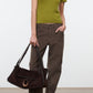 A woman is wearing the Staud Easy Cashmere Tee—a green, short-sleeved fitted top with a crew neckline—paired with brown wide-leg pants, black shoes, and holding a large brown suede handbag against a plain background.