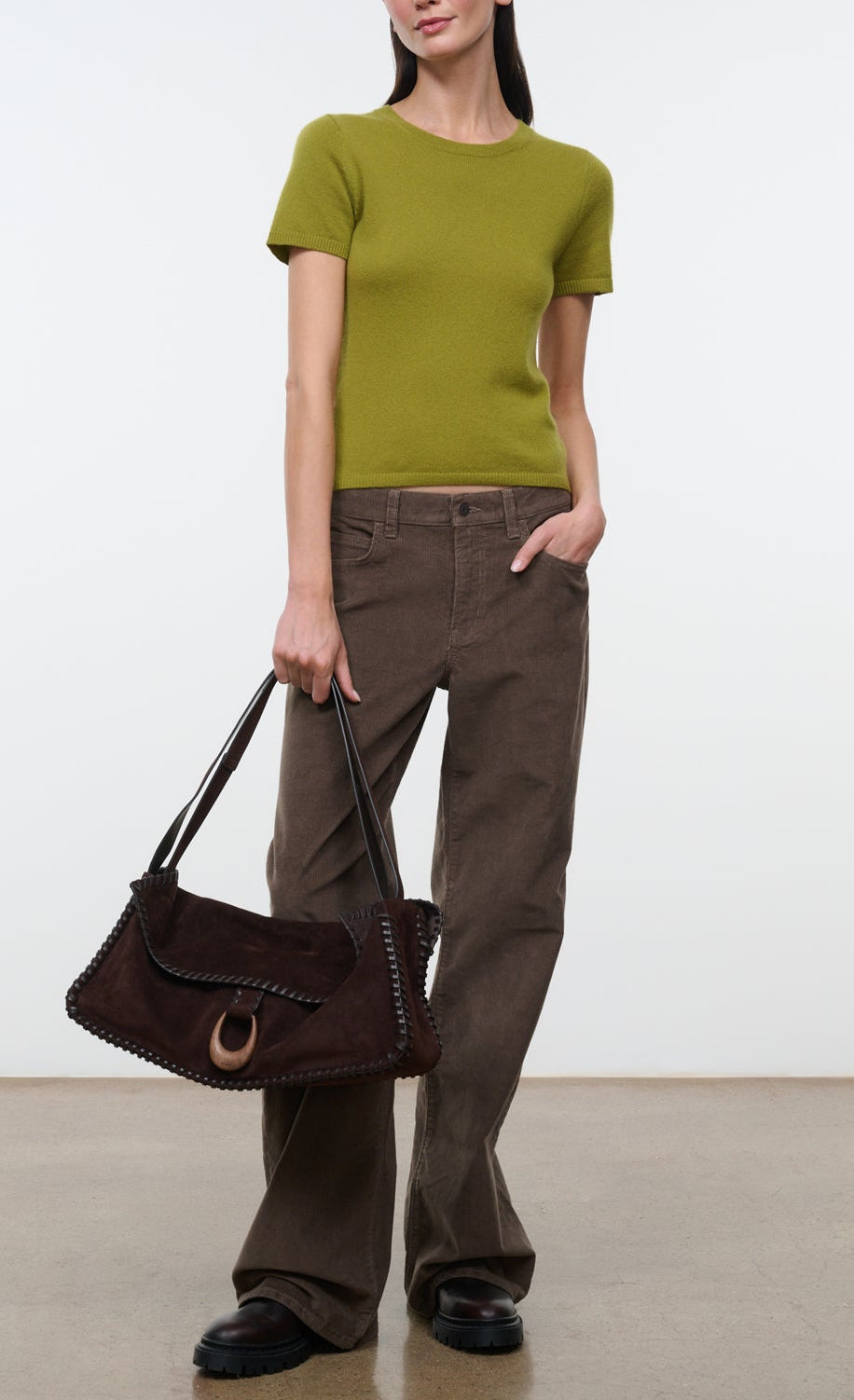 A woman is wearing the Staud Easy Cashmere Tee—a green, short-sleeved fitted top with a crew neckline—paired with brown wide-leg pants, black shoes, and holding a large brown suede handbag against a plain background.