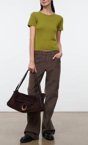 A woman is wearing the Staud Easy Cashmere Tee—a green, short-sleeved fitted top with a crew neckline—paired with brown wide-leg pants, black shoes, and holding a large brown suede handbag against a plain background.