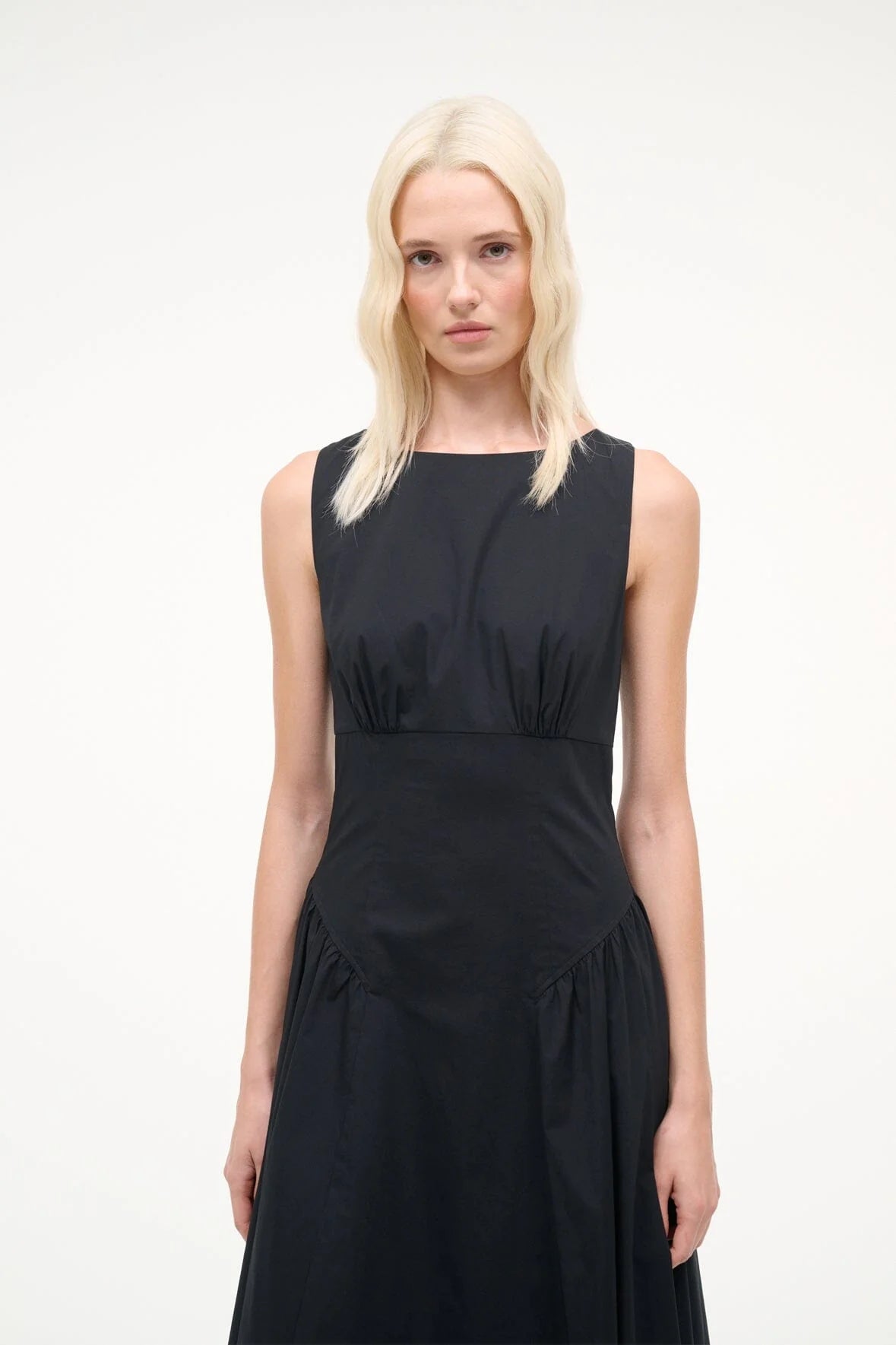 A blonde woman stands against a plain background, wearing the Gwen Dress by Staud—a sleeveless black A-line dress featuring a fitted waist and structured pleats that add subtle texture to its slightly flared skirt.