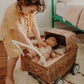 In a cozy bedroom, a child in a beige outfit enjoys imaginative play with a doll nestled in an Olli Ella Rattan Strolley on the rug.