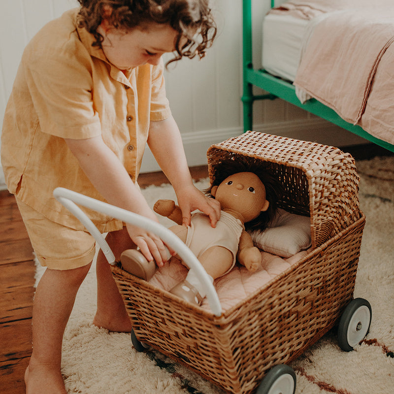 A child in a yellow shirt is putting an Olli Ella Rattan Strolley into a hand-woven rattan basket in a warmly lit bedroom.