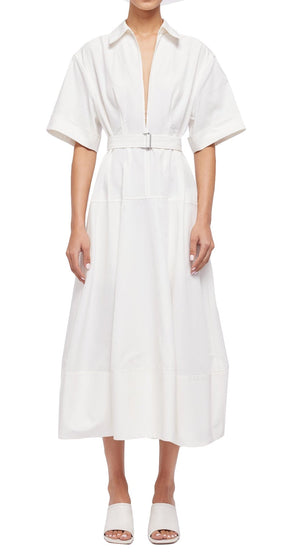 A person wearing the Simkhai Deanna Dress, featuring a belted waist, short sleeves, and an open collar, paired with white slip-on shoes, showcasing elevated everyday style with timeless craftsmanship.