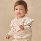A toddler dressed in the Quincy Mae Baby Girls’ and Girls’ Ruffle Fleece Sweatshirt in a cream shade holds a small sprig of dried flowers, standing against a plain beige background. This cozy sweatshirt features a charming ruffle V-detail, adding extra sweetness to her look.