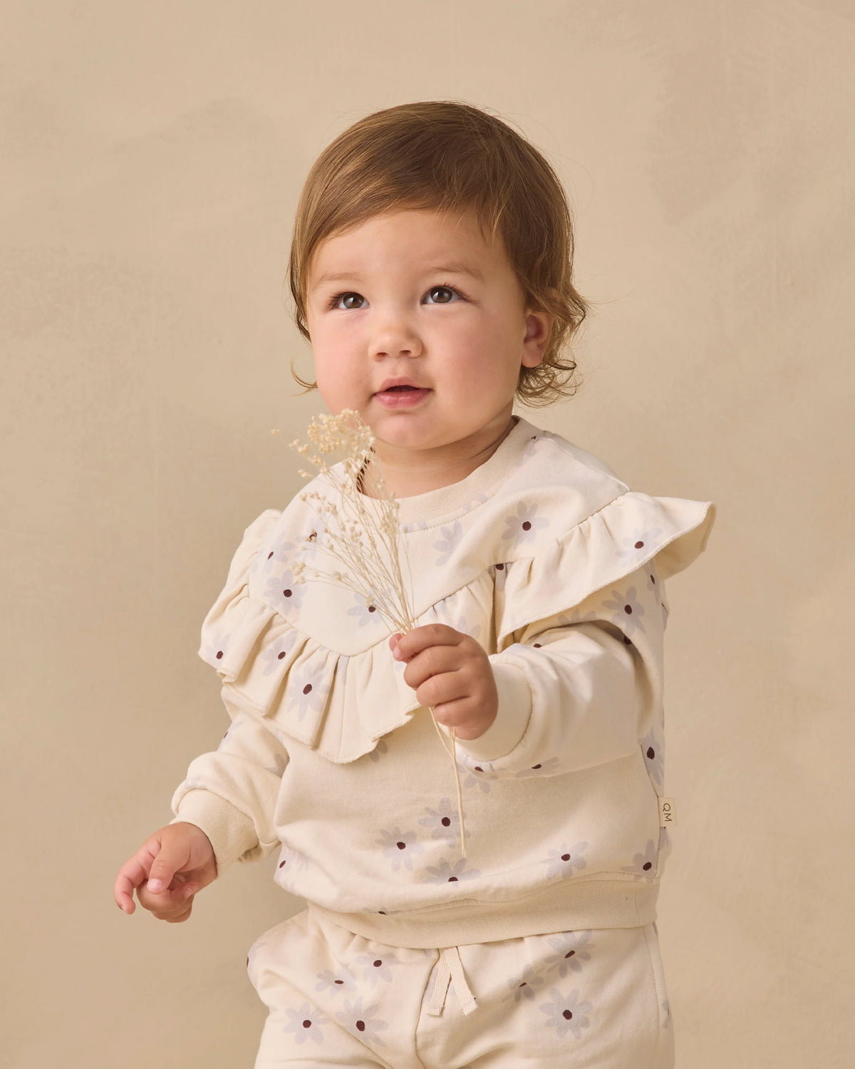 A toddler dressed in the Quincy Mae Baby Girls’ and Girls’ Ruffle Fleece Sweatshirt in a cream shade holds a small sprig of dried flowers, standing against a plain beige background. This cozy sweatshirt features a charming ruffle V-detail, adding extra sweetness to her look.