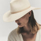 A person wearing the Janessa Leone Sherman Hat—a classic fedora with a wide brim—and a light v-neck blouse looks downward against a plain background, embodying timeless design.