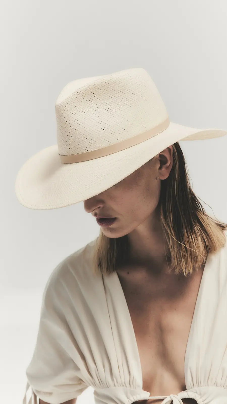 A person wearing the Janessa Leone Sherman Hat—a classic fedora with a wide brim—and a light v-neck blouse looks downward against a plain background, embodying timeless design.