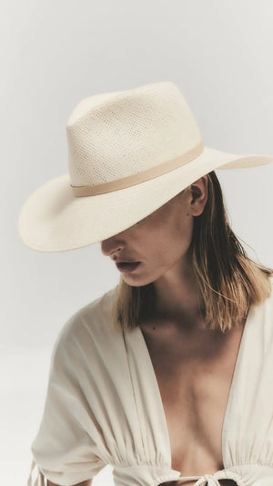 A person wearing the Janessa Leone Sherman Hat—a classic fedora with a wide brim—and a light v-neck blouse looks downward against a plain background, embodying timeless design.