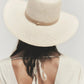 Individual with medium-length brown hair wears the Janessa Leone Sherman Hat, a wide-brimmed fedora, and an open-back cream dress, photographed from behind against a plain background.