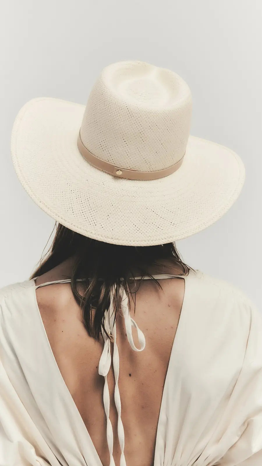 Individual with medium-length brown hair wears the Janessa Leone Sherman Hat, a wide-brimmed fedora, and an open-back cream dress, photographed from behind against a plain background.